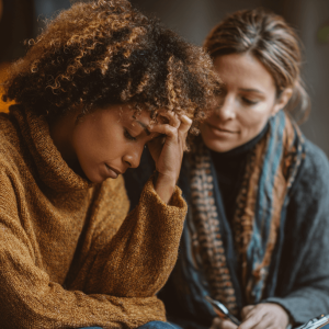 A supportive woman sits beside a distressed young woman during a compassionate, informal conversation — representing incidental counselling in the workplace.